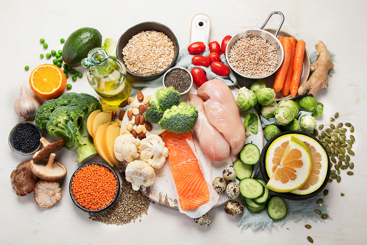 Assorted healthy foods including vegetables, grains, lean meats, seeds and citrus on a white table