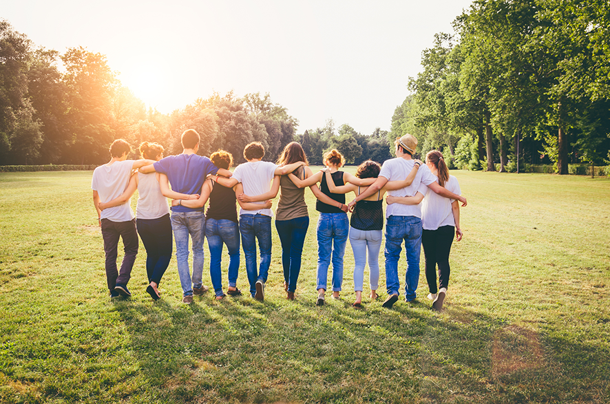 A group of 10 people link arms with each other from behind and walk through a park.