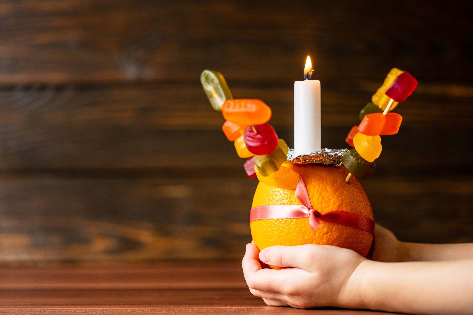 Two hands hold a Christingle orange, which is decorated with ribbon, sweets and a lit candle, against a wooden background