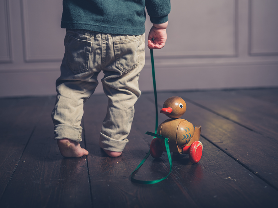 A small child pulls a wooden duck toy with wheels along, using a ribbon. The floor is wooden
