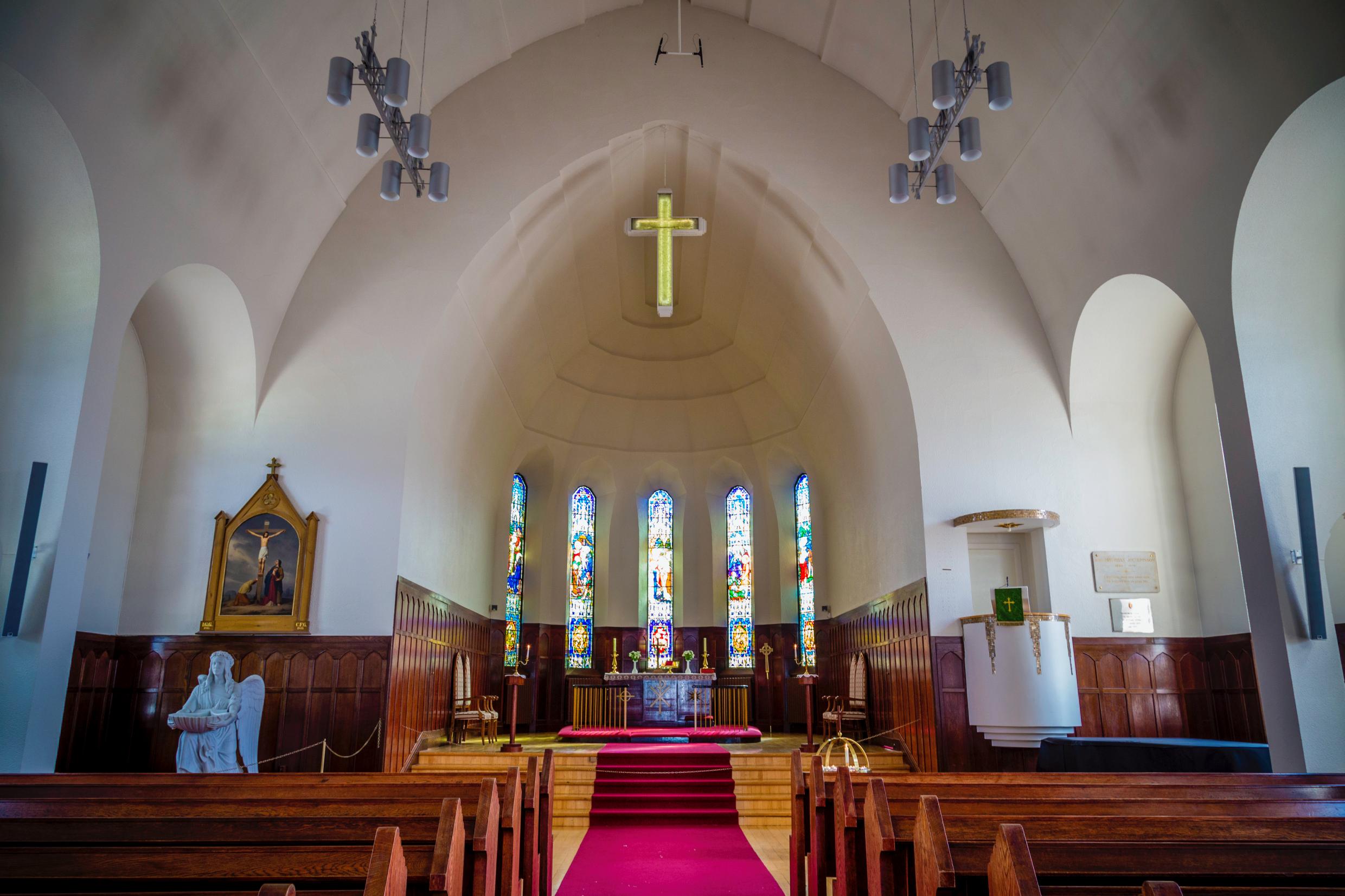 Interior of a church with wooden pews, stained glass windows, a raised altar, and a glowing cross above.