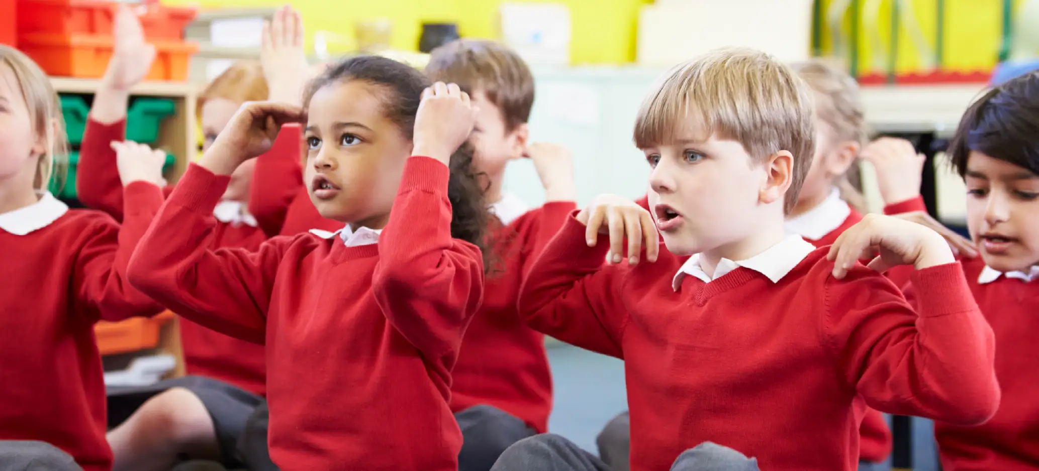 A group of primary school children in red school uniforms sit together in a classroom, raising their hands and touching their heads as they take part in an interactive lesson.