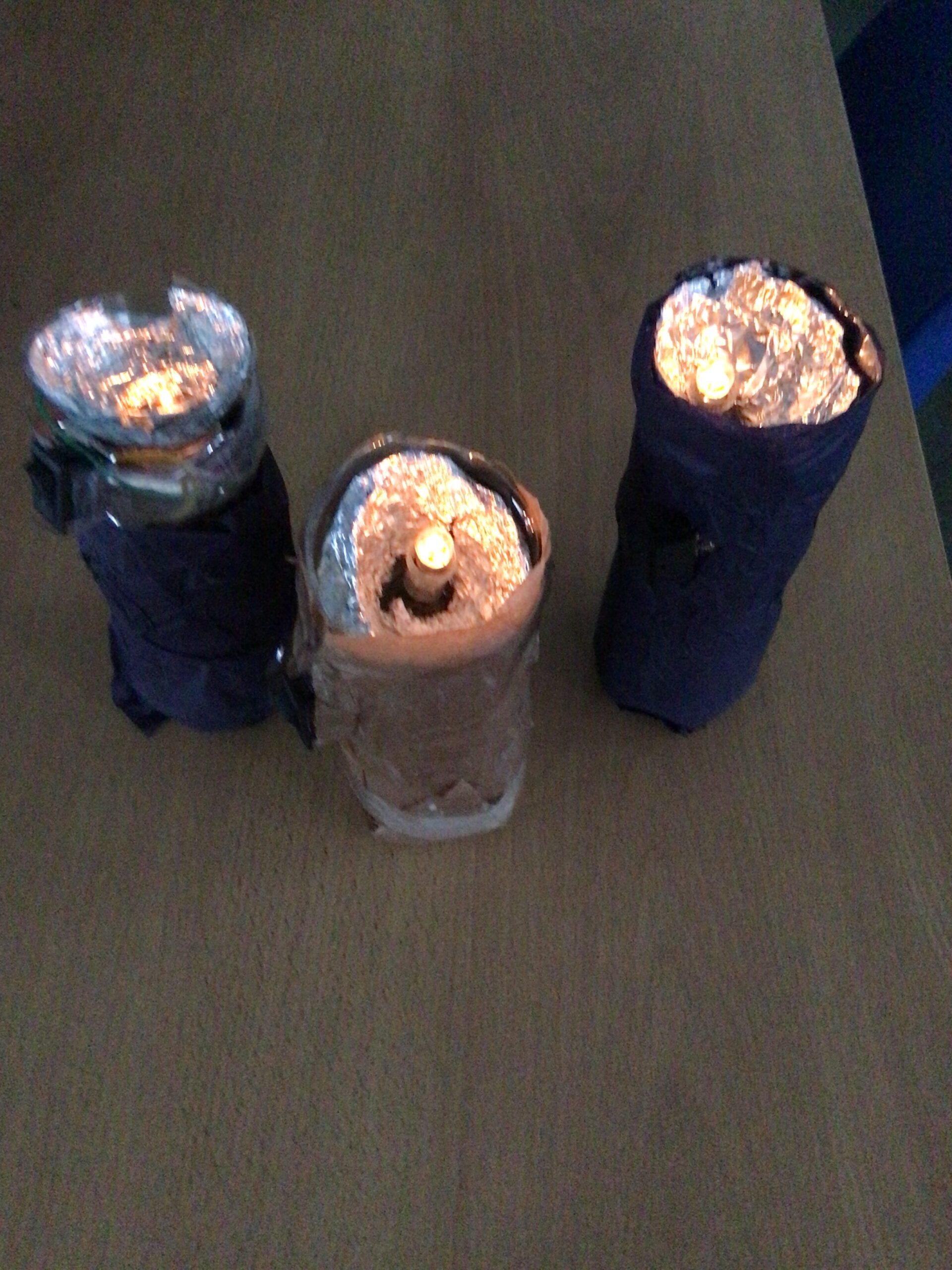 Three homemade torch models placed on a wooden desk. Each torch has a cylindrical design with outer coverings made of dark and light materials, such as paper or fabric. The tops are lined with reflective aluminum foil, and small light sources, possibly LEDs or candles, are visible emitting a warm glow. The arrangement forms a triangular grouping, and the background is a plain desk surface.