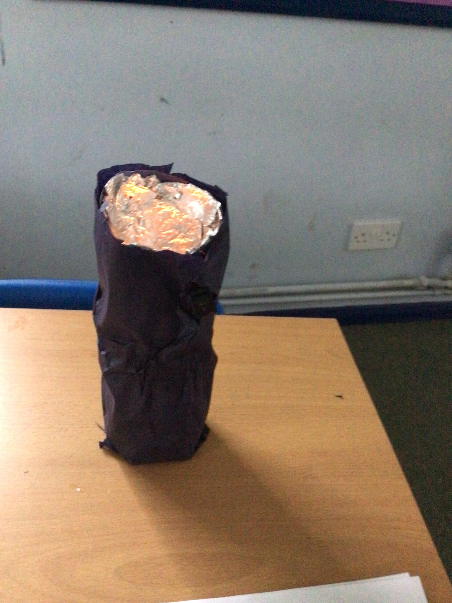 A homemade torch model placed on a wooden desk. It has a cylindrical shape wrapped in dark paper or fabric, with the top covered in crumpled aluminum foil, which appears to reflect light. The background includes part of a classroom wall, a power socket, and a blue chair