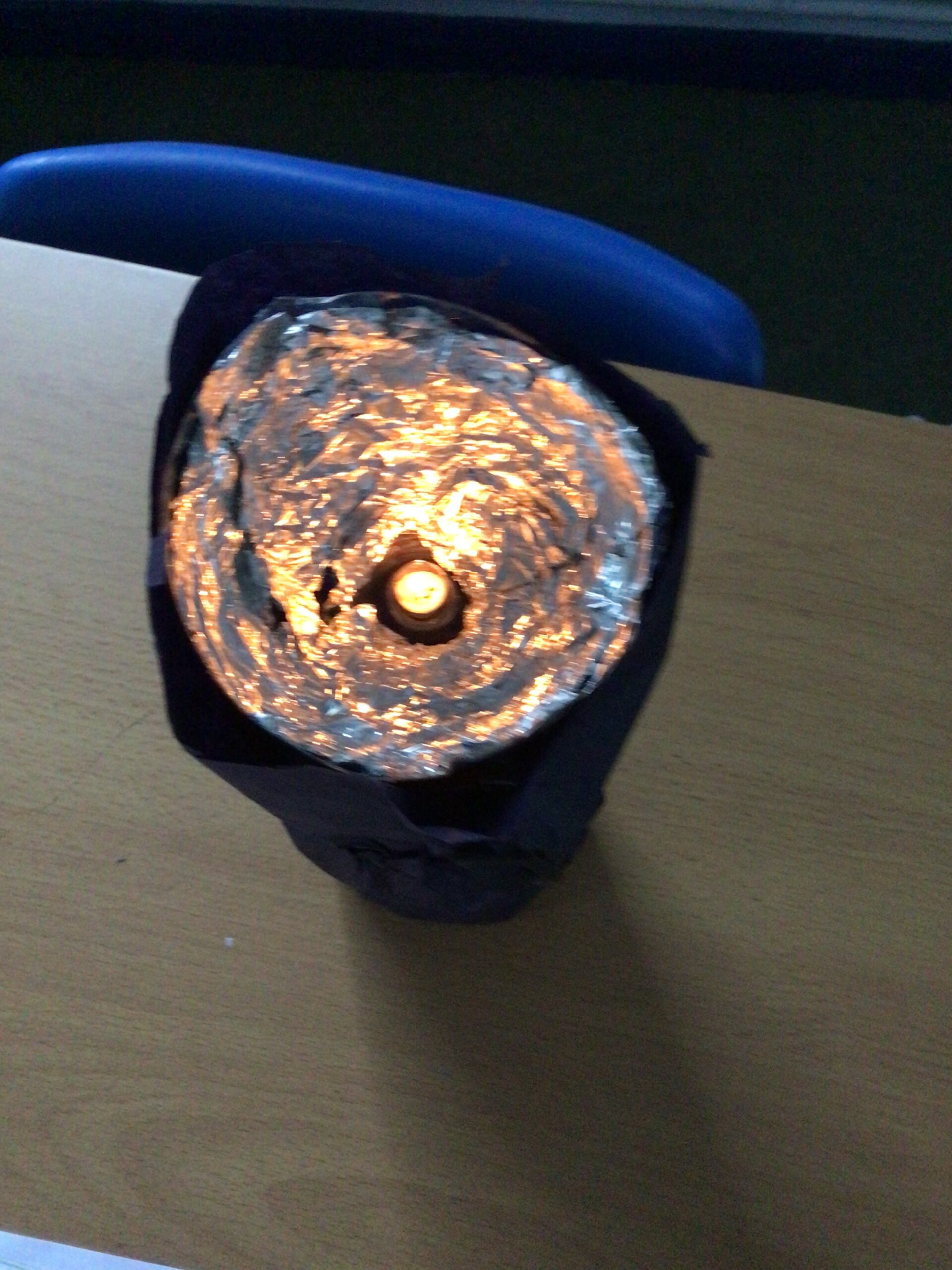 A close-up view of a homemade torch model on a wooden desk. The torch has a cylindrical shape wrapped in dark material, with the top covered in shiny, crumpled aluminum foil. A warm light source, possibly an LED or candle, is visible at the center, reflecting off the foil to create a glowing effect. In the background, part of a blue chair and a classroom setting are visible.