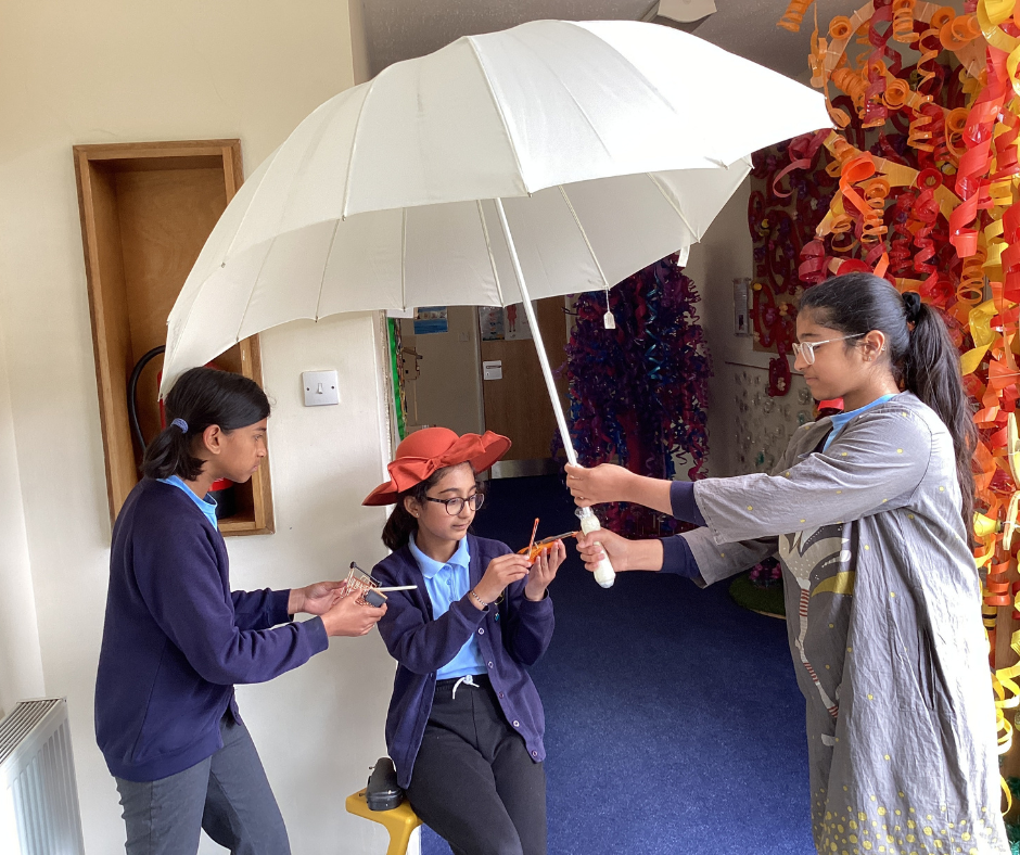 Three individuals indoors, one holding a large white umbrella over the other two. The seated person wears a red hat and holds an orange object, while another stands next to them holding a small rectangular item