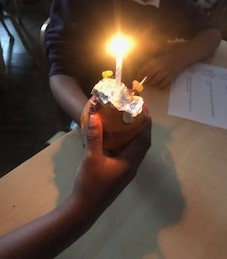 A photo of a person holding a completed and lit Christingle. The orange is decorated with a central candle wrapped in foil and cocktail sticks with sweets or fruits. The candle flame is glowing brightly, symbolizing light.